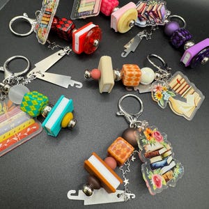 Może przedstawiać: Kolekcja breloków do kluczy z zawieszkami o tematyce książkowej. Zawieszki przedstawiają miniaturowe stosy książek, kolorowe koraliki i dekoracyjne akcenty. Każdy brelok ma srebrne zapięcie ułatwiające mocowanie.