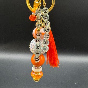 May include: A gold-colored keychain with a beaded design. The keychain features orange, white, and clear beads, with the text "UTHSCSLPPROGRAM" spelled out in clear letter beads. An orange tassel hangs from the bottom.