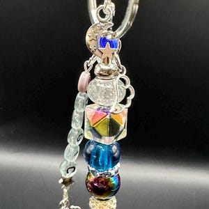 May include: A silver keychain with a variety of colorful beads and charms. The keychain features a moon and star charm, a blue star, a clear teapot charm, and a faceted iridescent cube. The bottom has a silver angel charm.