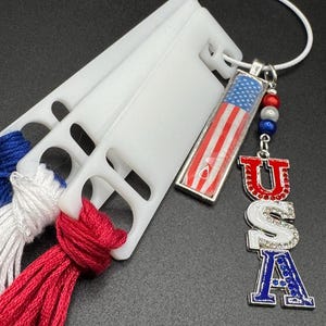 May include: A patriotic charm with a miniature American flag design, red, white, and blue beads, and a charm spelling out "USA" in red, white, and blue. The charm is attached to a white cord and is next to white plastic thread organizers with red, white, and blue thread.
