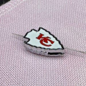 May include: A silver-toned pin featuring a white arrowhead design with the letters "KC" in red. The pin is set against a light pink fabric background, showcasing the pin's details.