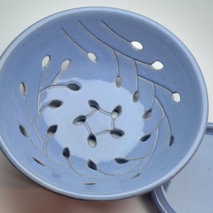 May include: A light blue ceramic bowl featuring a series of cut-out leaf-shaped patterns arranged in a swirling, radial design that originates from the center. The interior surface has a smooth, glossy finish with visible speckles throughout the glaze.