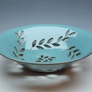May include: A shallow, round ceramic bowl with a glossy turquoise glaze. The bowl features intricate cut-out patterns of leafy vine branches and a central floral motif, revealing the open space through the clay body of the vessel.