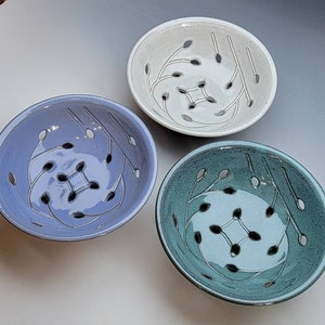 May include: Three ceramic colanders in different colors: white, light blue, and teal. Each colander has a unique design of stylized leaves and holes for draining.