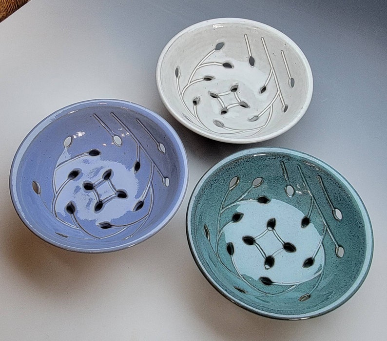 May include: Three ceramic colanders in different colors: white, light blue, and teal. Each colander has a unique design of stylized leaves and holes for draining.