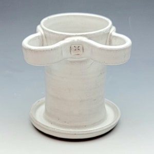 May include: White ceramic mug with two handles and a small plate base. The mug has a unique design with a raised band around the middle and a small square embossed on the front.