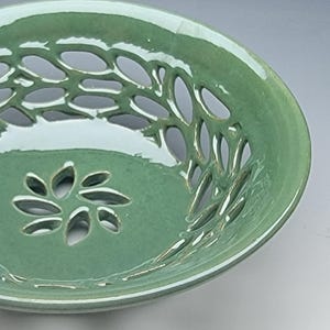 May include: A sage green ceramic bowl featuring a glossy finish and an intricate openwork lattice pattern along the sides. The base of the bowl displays a cutout design shaped like a six-petaled flower, creating a decorative and functional vessel.