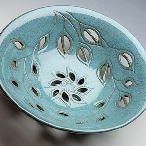 May include: A round ceramic bowl with a glossy teal glaze, featuring intricate cut-out leaf patterns along the sides and a central flower-shaped cut-out design. The interior surface displays a textured finish with visible carved details.