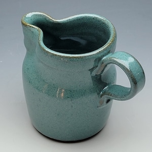 May include: A handmade turquoise ceramic creamer with a curved spout and a rounded handle. The creamer has a textured surface and a slightly flared rim with a brown edge. The creamer is approximately 4 inches tall.