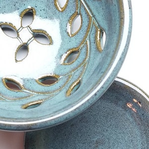 May include: A teal ceramic bowl with a white interior and gold leaf design. The bowl has a decorative cut-out pattern of leaves. The bowl is sitting on a smaller teal ceramic plate.