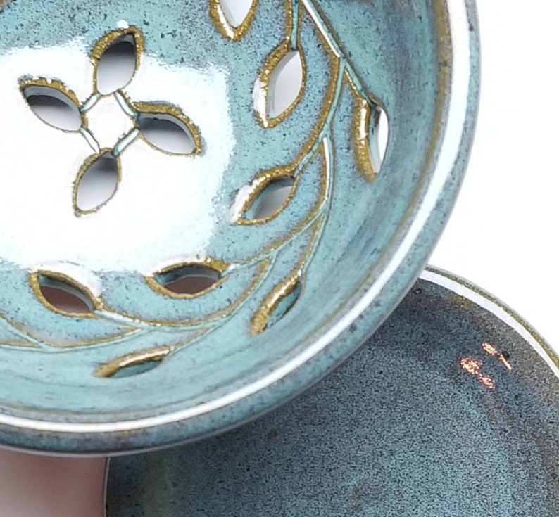 May include: A teal ceramic bowl with a white interior and gold leaf design. The bowl has a decorative cut-out pattern of leaves. The bowl is sitting on a smaller teal ceramic plate.