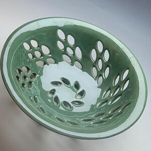 May include: A round ceramic bowl with a glossy sage green glaze on the exterior and interior rim. The bowl features a series of evenly spaced, oval-shaped cutouts arranged in a circular pattern around the sides and base of the vessel.