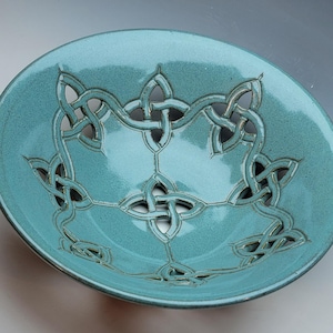 May include: A round, teal-colored ceramic bowl featuring an intricate, openwork cutout design of interconnected Celtic knots. The glossy, smooth glaze covers the entire surface, highlighting the detailed, geometric patterns carved into the clay.