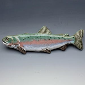 May include: A ceramic trout figurine featuring a glossy finish with a green back, a pink lateral stripe, and a white underbelly. The fish has dark spots along its side, detailed fins, and a textured tail, capturing the appearance of a rainbow trout.