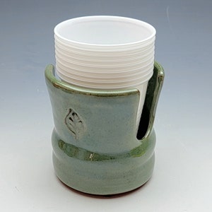 May include: A green ceramic cup holder with a leaf design, holding a stack of white plastic cups.