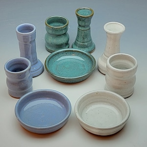 May include: A set of eight ceramic candle holders in various shades of blue, white, and green. The holders are arranged in a circle on a white surface. Some of the holders have a textured finish.