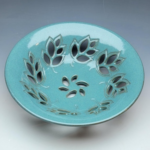 May include: A round, teal-colored ceramic bowl featuring a glossy finish and intricate cut-out patterns. The design includes a central floral-like motif surrounded by a ring of stylized leaf shapes, all with dark, defined edges.