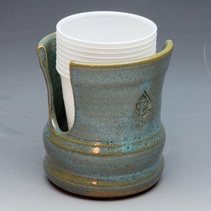 May include: A teal green ceramic cup holder with a leaf design. The holder is filled with white disposable cups.