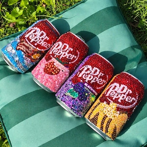 May include: Four aluminium soda cans decorated with colourful rhinestones. Each can features the Dr Pepper logo and specific flavour text including Creamy Coconut, Strawberry Cream, Blackberry, and Cream Soda, all covered in sparkling crystal embellishments.
