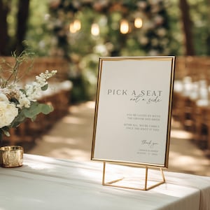 May include: A gold-framed sign with the text "PICK A SEAT not a side" in black script. The sign also includes the text "YOU'RE LOVED BY BOTH THE GROOM AND BRIDE" and "AFTER ALL, WE'RE FAMILY ONCE THE KNOT IT TIED". The sign is on a gold stand.