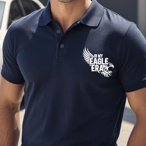 May include: Navy blue polo shirt with a white graphic of an eagle and the text "IN MY EAGLE ERA." The shirt has a classic collar and buttoned placket. The shirt is made of a soft, comfortable material.