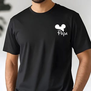 May include: Black t-shirt with a white graphic of Mickey Mouse ears and the word "PAPA" in a stylized font. The shirt is short-sleeved and made of a soft material.