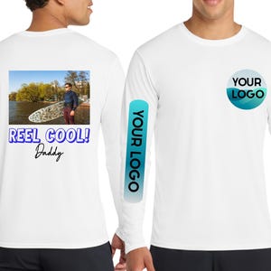 May include: White long-sleeve shirt with a graphic on the back that says "Reel Cool! Daddy" below a photo of a person holding a surfboard. The shirt also features a logo on the sleeve and chest.