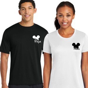 May include: Black and white t-shirts with a Mickey Mouse head design. The black shirt has the word "Papa" and the white shirt has the word "Minnie". Both shirts are short-sleeved and worn by two people.