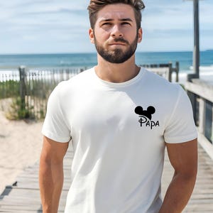 May include: White t-shirt with a black Mickey Mouse head and the word "PAPA" printed below it. The t-shirt is short-sleeved and has a crew neck. The background shows a beach scene with a wooden walkway and the ocean.