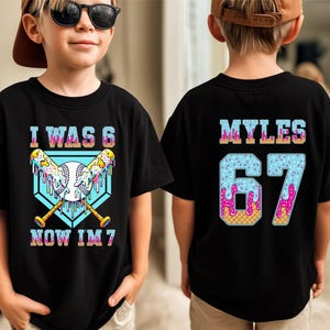 May include: Black t-shirt with a baseball-themed graphic. The front features the text "I WAS 6 NOW IM 7" with baseball bats and a baseball. The back displays the name "MYLES" and the number "67" in a colorful, ice cream-themed design.