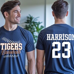 May include: Navy blue baseball t-shirt with "TIGERS Baseball" in orange and white lettering on the front. The back of the shirt displays "HARRISON 23" in white. The shirt is worn by a person.