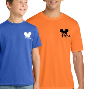 May include: Two short-sleeved t-shirts, one blue and one orange. The blue shirt has a white Mickey Mouse head design with the word "Mickey" below it. The orange shirt has a black Mickey Mouse head design with the word "Papa" below it.