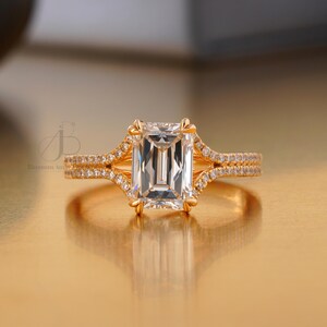 Könnte beinhalten: Ein goldener Verlobungsring mit einem großen, rechteckigen Diamanten im Smaragdschliff als Mittelpunkt. Das Band hat ein geteiltes Schaftdesign mit kleineren Akzentdiamanten. Der Ring ist vor einem warmen, goldenen Hintergrund platziert.