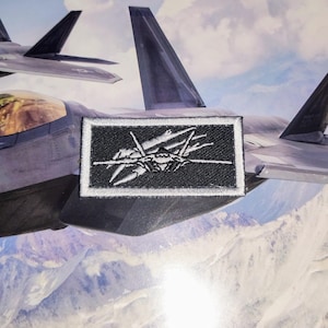 May include: Embroidered patch featuring a stylized jet fighter design in white on a black rectangular background. The patch is set against a backdrop of two fighter jets in flight over a mountain range.