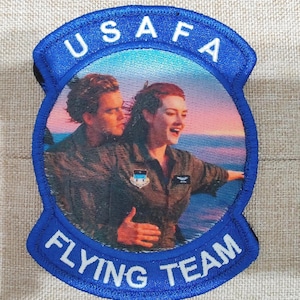 May include: A circular embroidered patch with a blue border featuring the text USAFA at the top and FLYING TEAM at the bottom. The centre displays a printed image of two individuals in flight suits standing against a sunset ocean background.