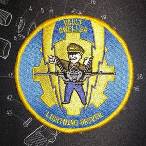 May include: Embroidered patch with a blue and yellow design. The patch features a cartoon character in a pilot's uniform, with the text "VAULT DWELLER" and "LIGHTNING DRIVER". The patch has a yellow border.