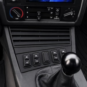 May include: A detailed shot of a car's interior, focusing on the black dashboard. It features air vents, control buttons, and a gear stick. The image has a dark, streamlined aesthetic, highlighting the car's design and functionality.