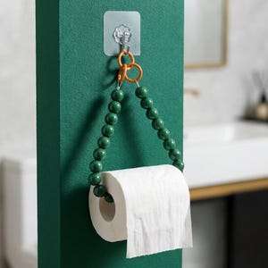 Könnte beinhalten: Ein dekorativer Toilettenpapierhalter mit einer Rolle weißem Papier. Der Halter besteht aus einer Kette grüner Perlen, die an einem orangefarbenen Ring aufgehängt sind und mit einem transparenten Klebehaken an der Wand befestigt sind. An einer dunkelgrünen Wand.