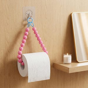 Puede incluir: Portarrollos de papel higiénico con cuentas rosas y un detalle azul, colgado de un gancho adhesivo transparente en una pared de madera. El soporte sostiene un rollo de papel higiénico blanco. Una vela y un espejo están en el fondo.