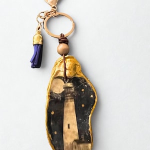 May include: A gold-tone keychain with an oyster shell pendant. The pendant depicts a lighthouse scene against a starry night sky. A small blue tassel and wooden bead are attached to the keychain.