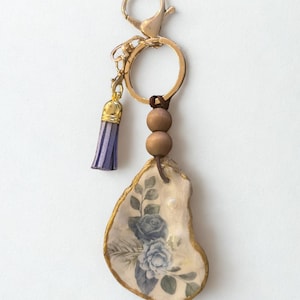 May include: A decorative keychain with an oyster shell pendant. The shell is painted with a blue floral design and has a gold edge. It's attached to a gold keyring with a purple tassel and wooden beads. The overall design is elegant.