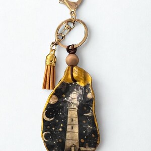 May include: A gold-coloured keychain with a painted, irregular-shaped pendant. The pendant depicts a lighthouse scene against a starry night sky, with crescent moons and stars. A small tan tassel hangs from the keychain.
