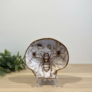 May include: A decorative seashell dish with a detailed illustration of a bee and floral accents. The dish has a gold-painted rim and is displayed on a clear stand. The bee design is in shades of brown and gold.