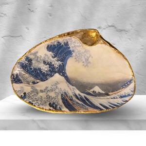 May include: A large, decorative seashell with a gold-painted rim. The interior features a detailed print of Hokusai's "The Great Wave off Kanagawa" in shades of blue, white, and grey. The shell is displayed on a white surface.