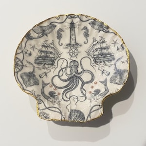 May include: A decorative seashell-shaped dish with a nautical theme. The white dish features a black and white illustration of an octopus, ships, lighthouse, seahorses, and mermaids. The rim is trimmed with gold.