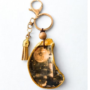 May include: Gold-toned keychain featuring an oyster shell pendant with a lighthouse and compass design. The shell is accented with gold paint and a small wooden bead. A gold tassel hangs from the chain.