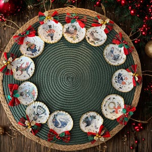 Puede incluir: Un conjunto de adornos navideños con forma de concha. Cada adorno presenta una ilustración única con ribete dorado, adornado con lazos escoceses rojos y verdes. Los adornos están dispuestos sobre una estera verde tejida, rodeados de decoraciones navideñas.