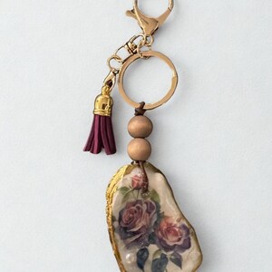 May include: A gold-coloured keyring with a burgundy tassel and wooden beads. The main feature is an oyster shell pendant with a floral rose design and gold edging. The keyring is attached to a clasp and a key ring.