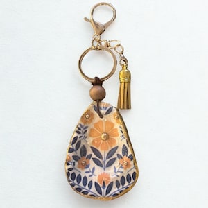 May include: A decorative keychain with a gold-coloured clasp, ring, and tassel. The pendant is teardrop-shaped, featuring a floral design in orange, navy, and white, with gold trim. A wooden bead accents the design.