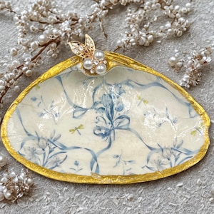 May include: A decorative seashell trinket dish with a gold-painted rim and a cream interior with a blue floral pattern. A gold and pearl embellishment adorns the top. The dish is surrounded by pearl accents. The dish is approximately 15 cm in length.